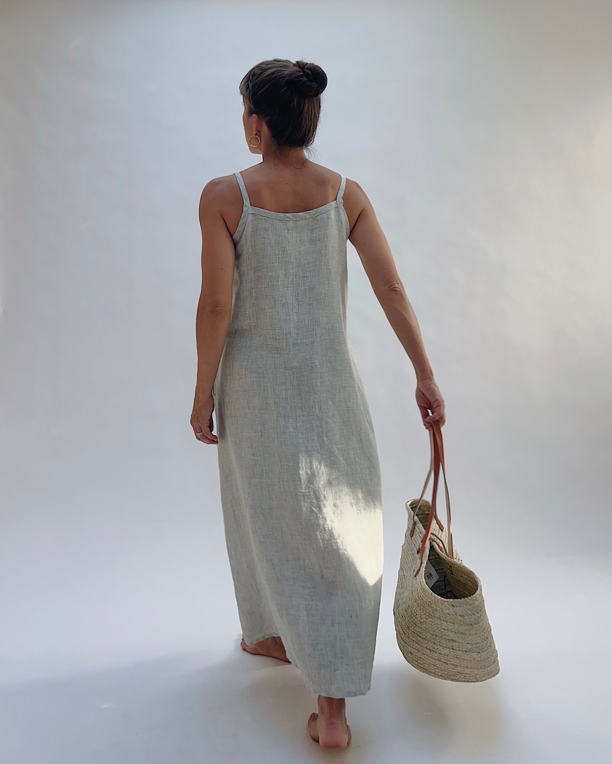 Model wearing the Yuvita Button Front Dress in Sage Pinstripe Stripe, a flowy linen maxi dress with button-front detail and spaghetti straps holding a woven tote bag and seen walking away from the camera