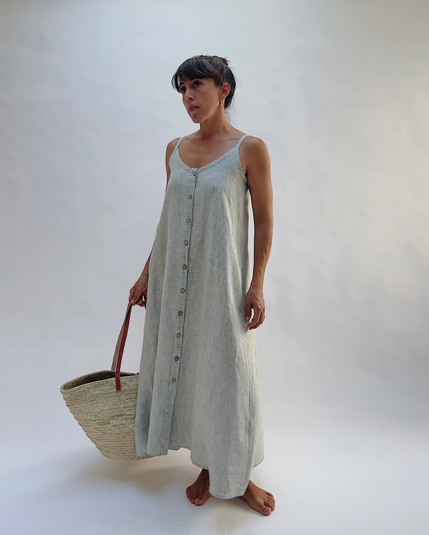 Model wearing the Yuvita Button Front Dress in Sage Pinstripe Stripe, a flowy linen maxi dress with button-front detail and spaghetti straps holding a woven tote bag
