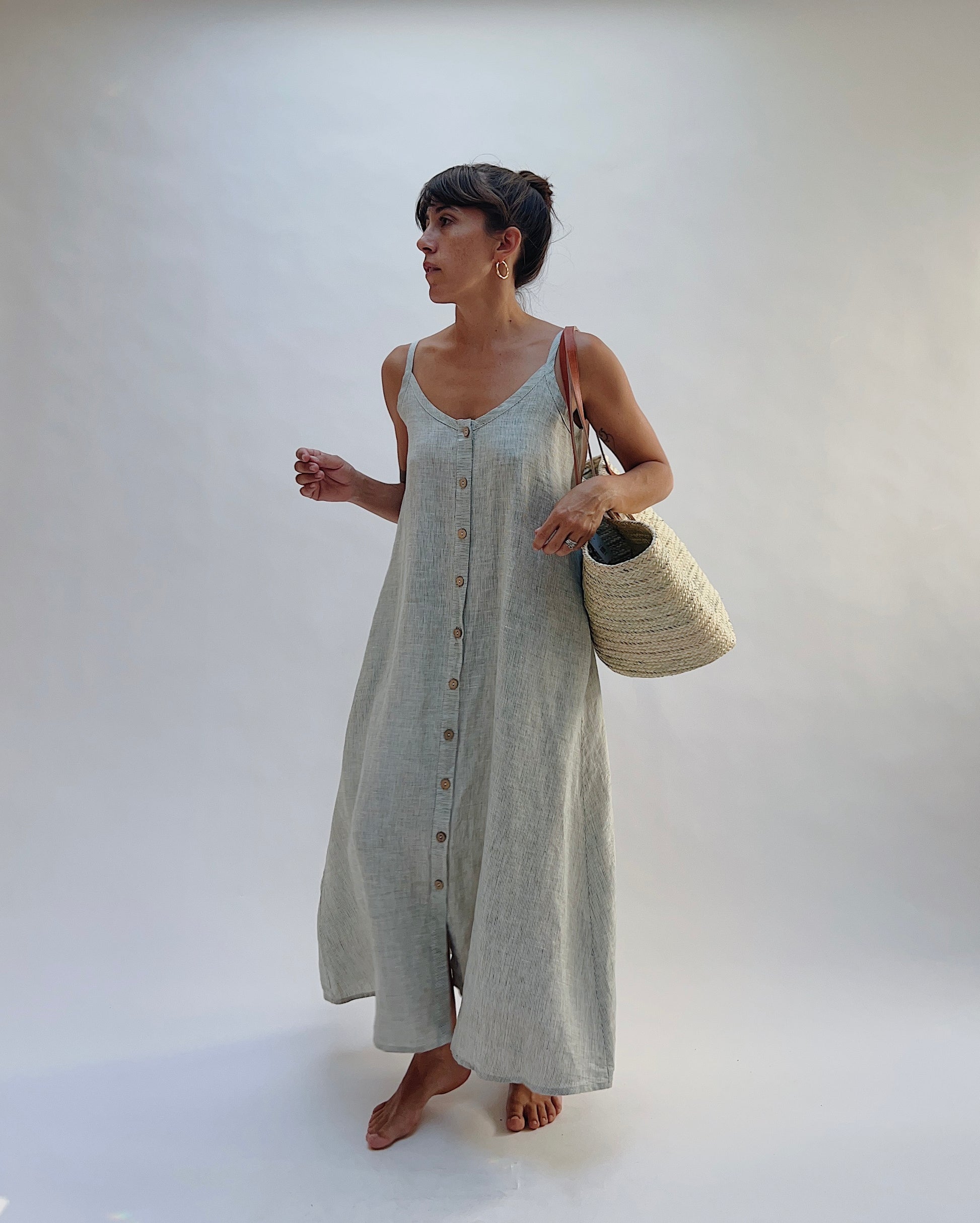 Model wearing the Yuvita Button Front Dress in Sage Pinstripe Stripe, a flowy linen maxi dress with button-front detail and spaghetti straps wearing a woven tote bag over one shoulder