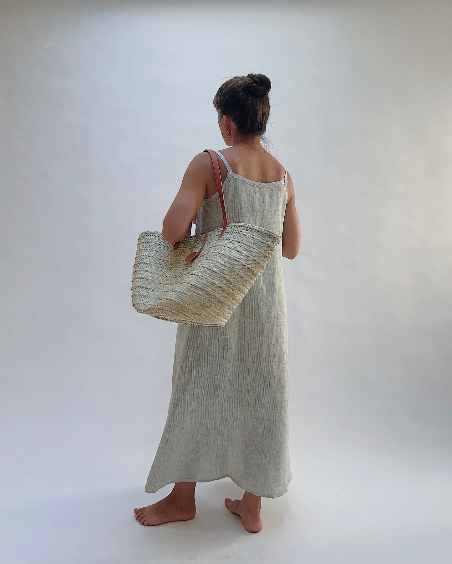 Model wearing the Yuvita Button Front Dress in Sage Pinstripe Stripe, a flowy linen maxi dress with button-front detail and spaghetti straps wearing a woven tote bag over one shoulder as seen from behind