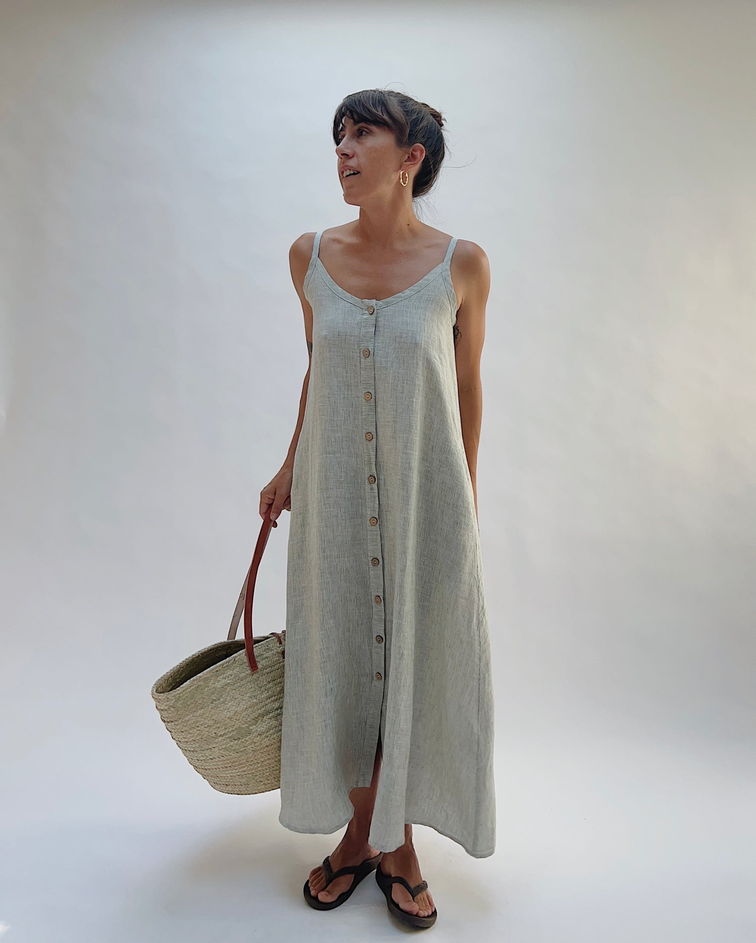 Model wearing the Yuvita Button Front Dress in Sage Pinstripe Stripe, a flowy linen maxi dress with button-front detail and spaghetti straps, styled with sandals and a woven tote bag