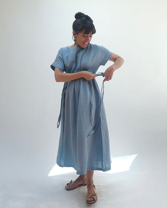 Woman with dark hair in a bun wearing a light blue linen dress and green dangle earrings standing against a white background tying the dress strings at one side of her body.