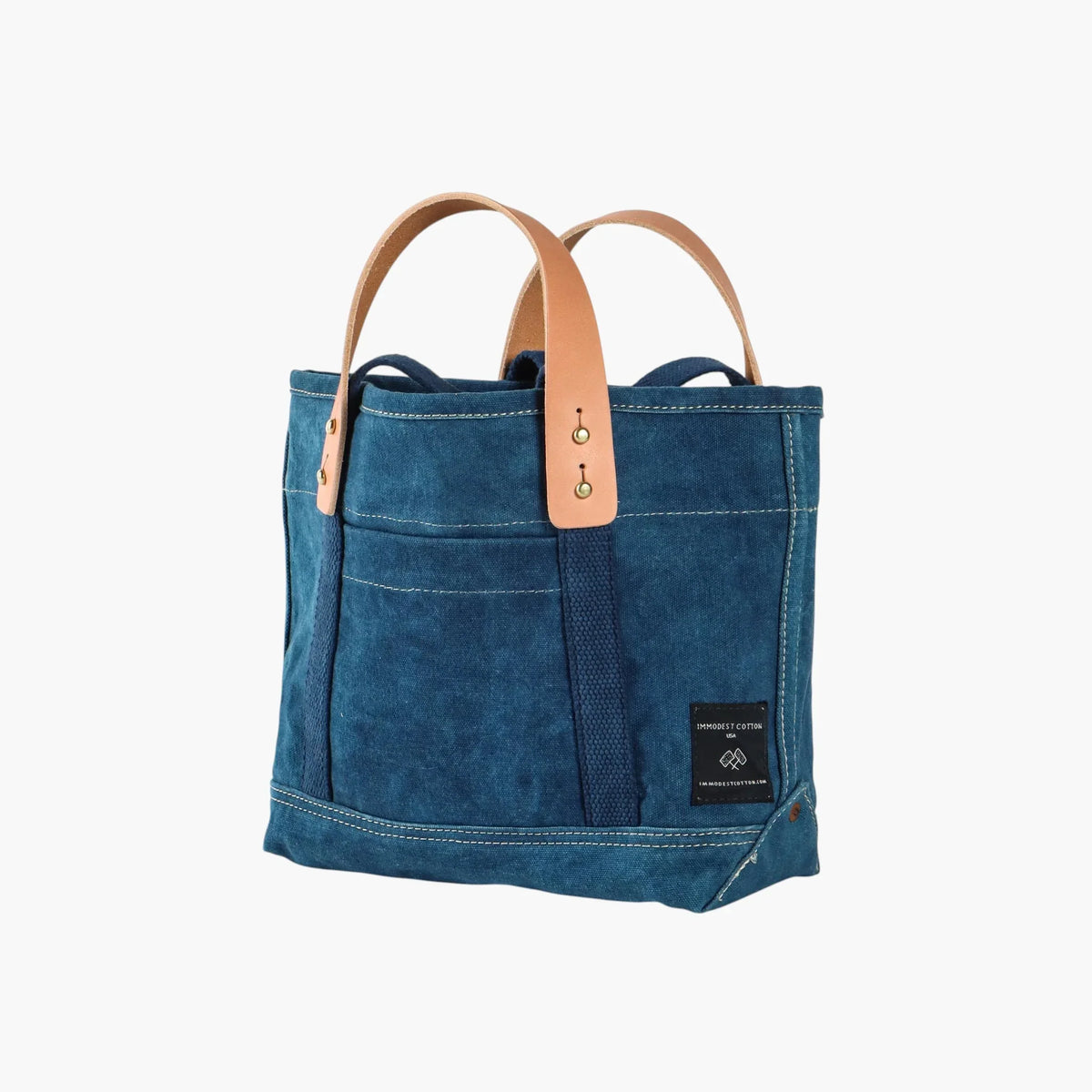 Angled view of the indigo Lunch Tote showing natural leather handles, the front pocket, and reinforced bottom corner.
