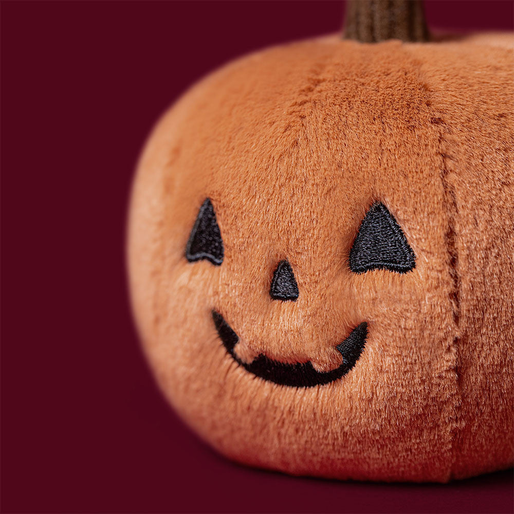 Close-up of the Jellycat Ooky Jack O Lantern face against a deep red background, showing the embroidered triangular eyes, nose, and grinning jack-o-lantern mouth on soft orange plush