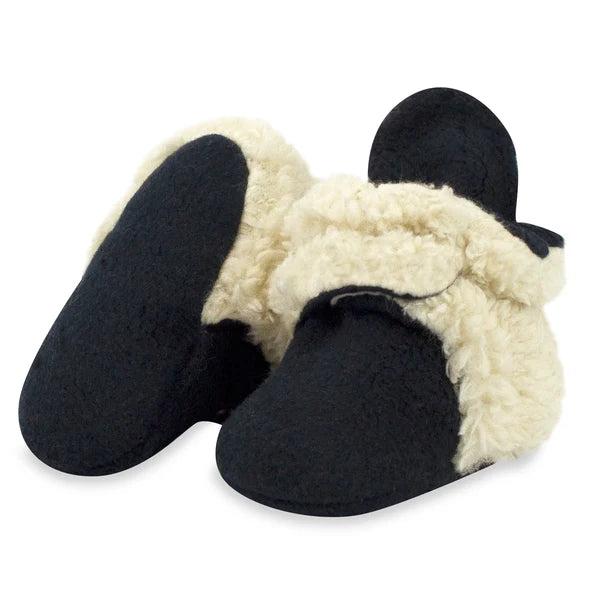 Black baby booties with cream faux fur trim, one tilted to show the plain fabric sole used in sizes 3M and 6M.
