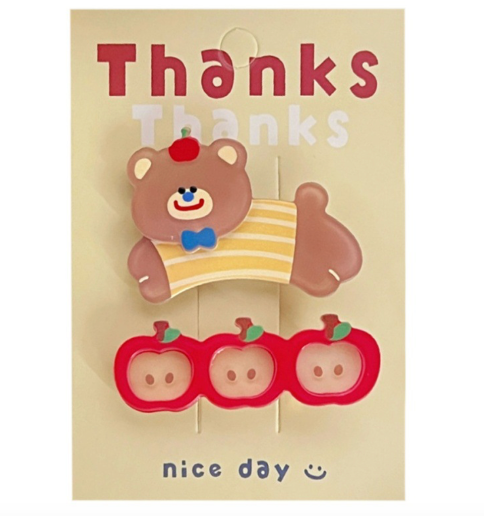A set of two hair clips with a bear and apple design shown displayed on a paper card