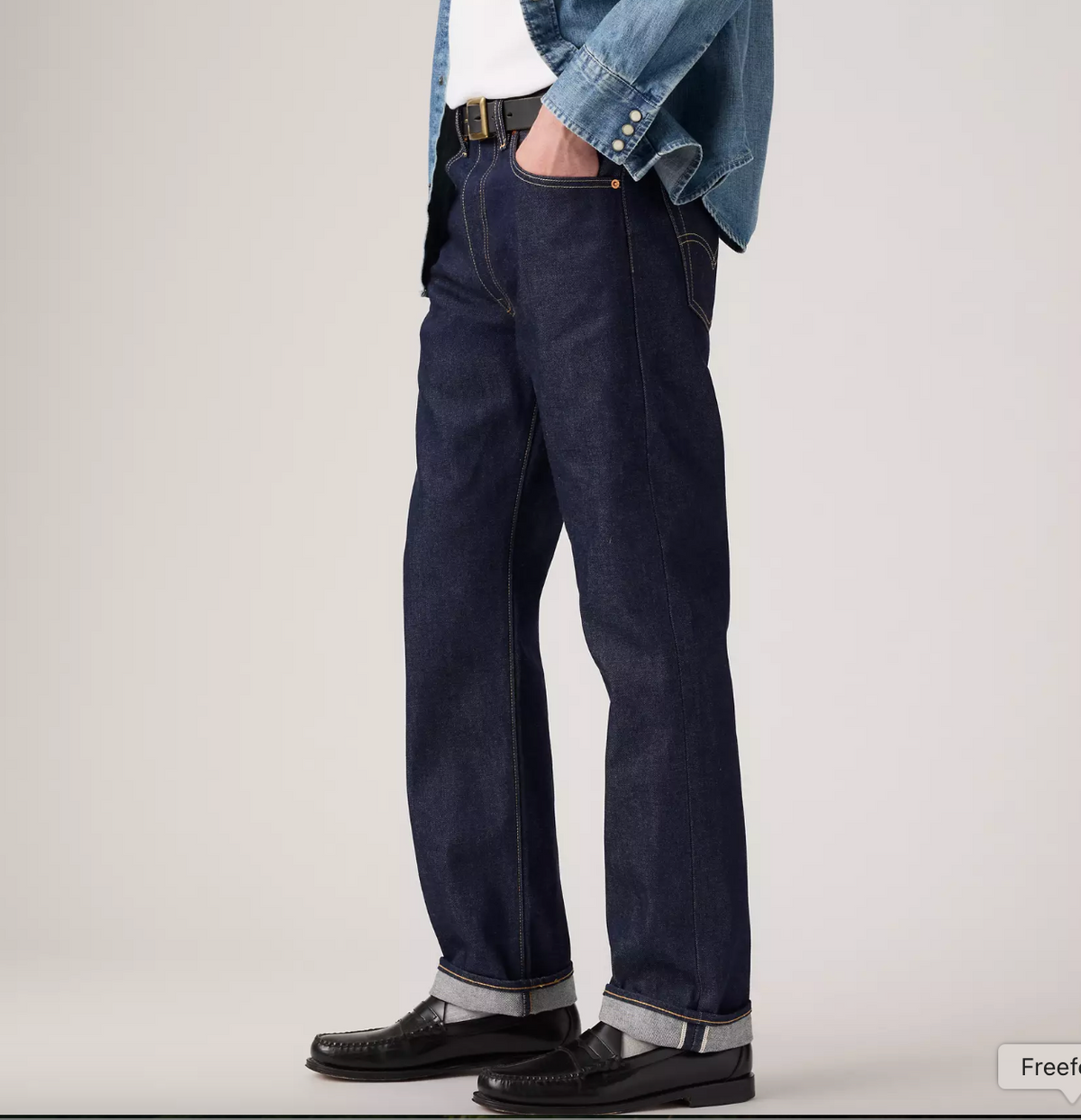 A person wearing dark indigo straight-leg jeans with a white shirt, a light blue denim jacket, and black leather shoes, shown from the waist down against a plain light gray studio background.