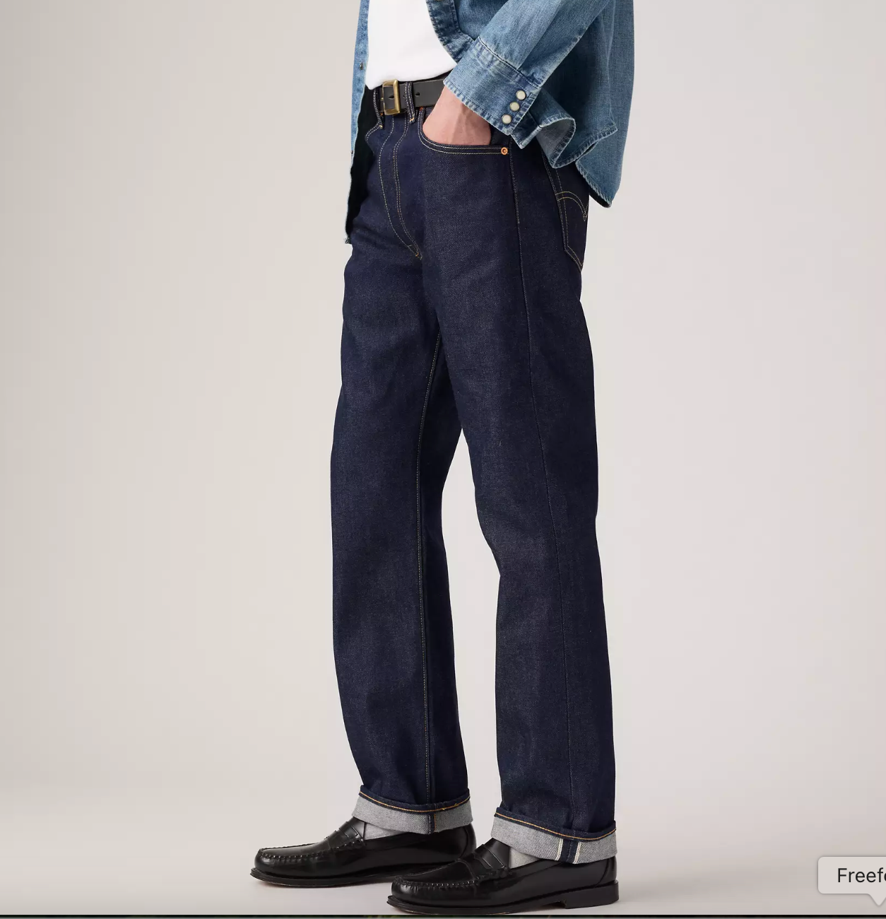 A person wearing dark indigo straight-leg jeans with a white shirt, a light blue denim jacket, and black leather shoes, shown from the waist down against a plain light gray studio background.