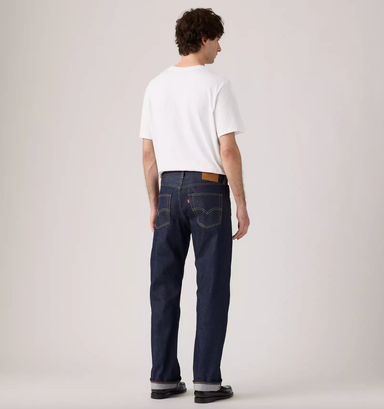 A man is shown from behind wearing a white t-shirt, dark blue Levi's jeans, and black shoes against a plain light gray studio background.