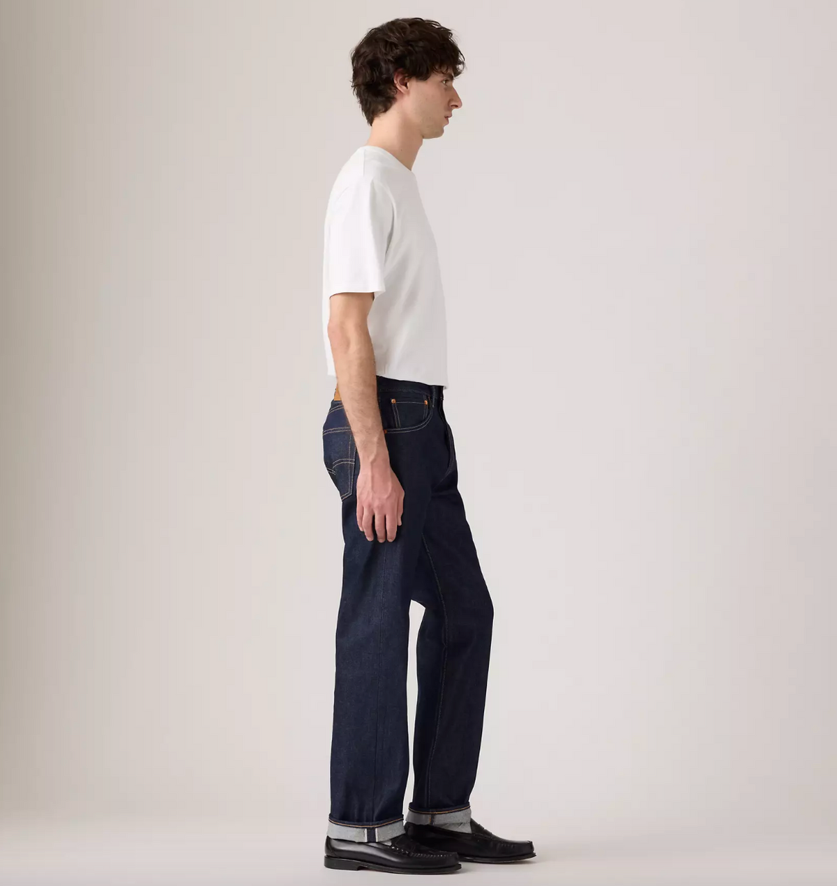 A man shown in profile wearing a white short-sleeve crewneck t-shirt, dark blue straight-leg jeans, and black leather shoes, standing against a plain light gray studio background.
