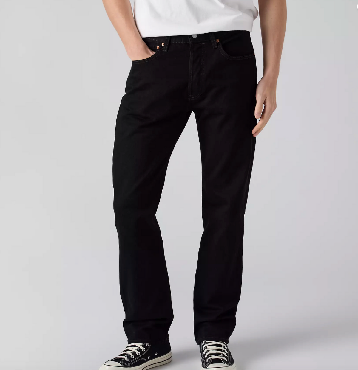 A person wearing black straight-leg jeans, a white t-shirt, and black canvas sneakers, shown from the waist down against a light gray studio background.