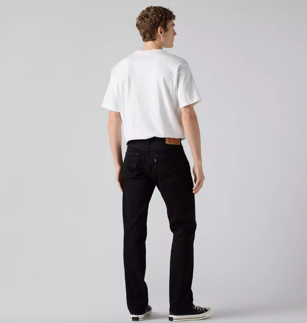 A man is shown from behind wearing a white t-shirt, black jeans, and black canvas sneakers against a light gray studio background.