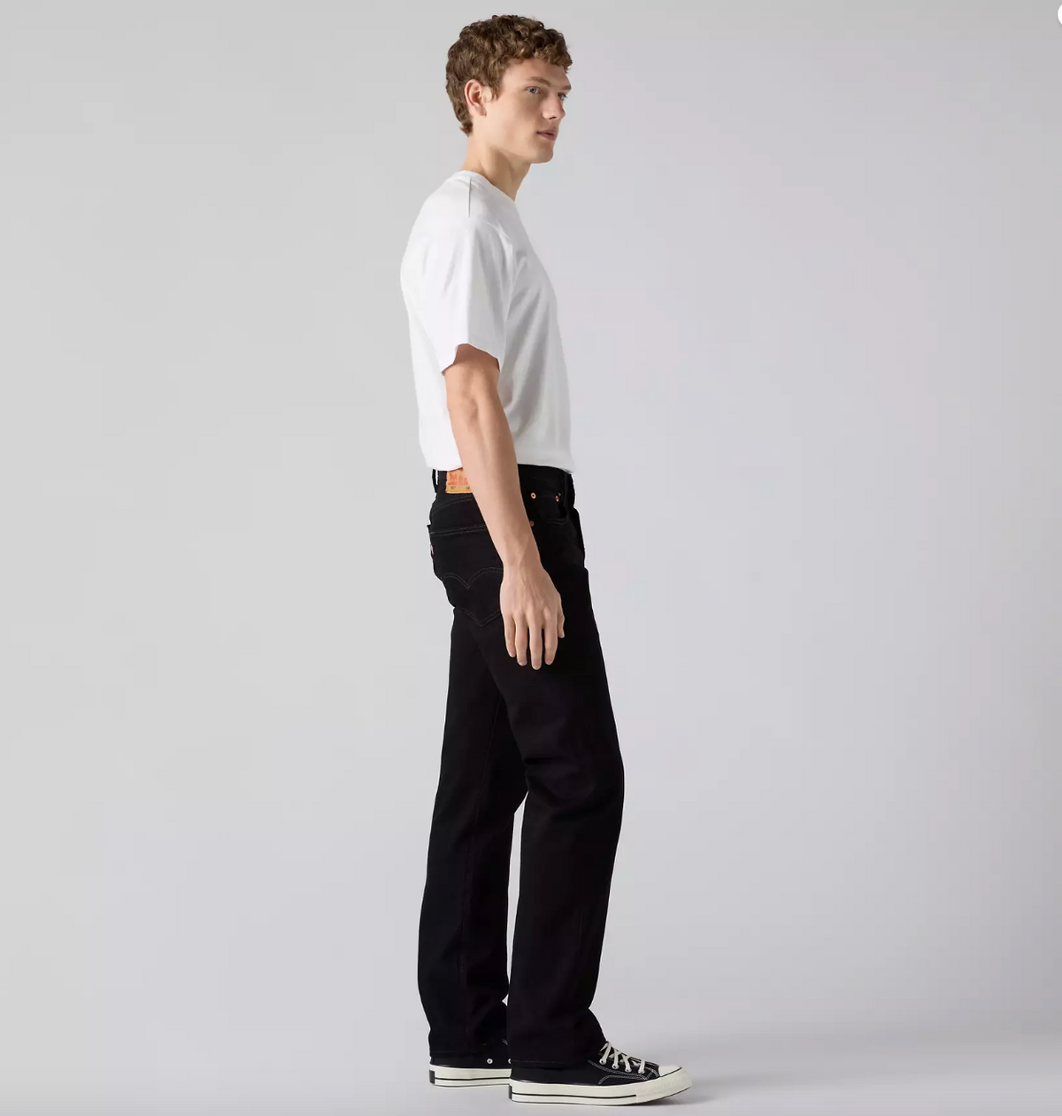 A man in a white t-shirt and black jeans is shown in profile against a light gray studio background.