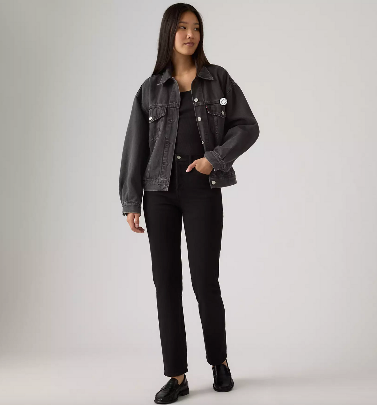 A model stands against a light gray studio background wearing a dark gray washed denim jacket over a black top and black pants, with black shoes.