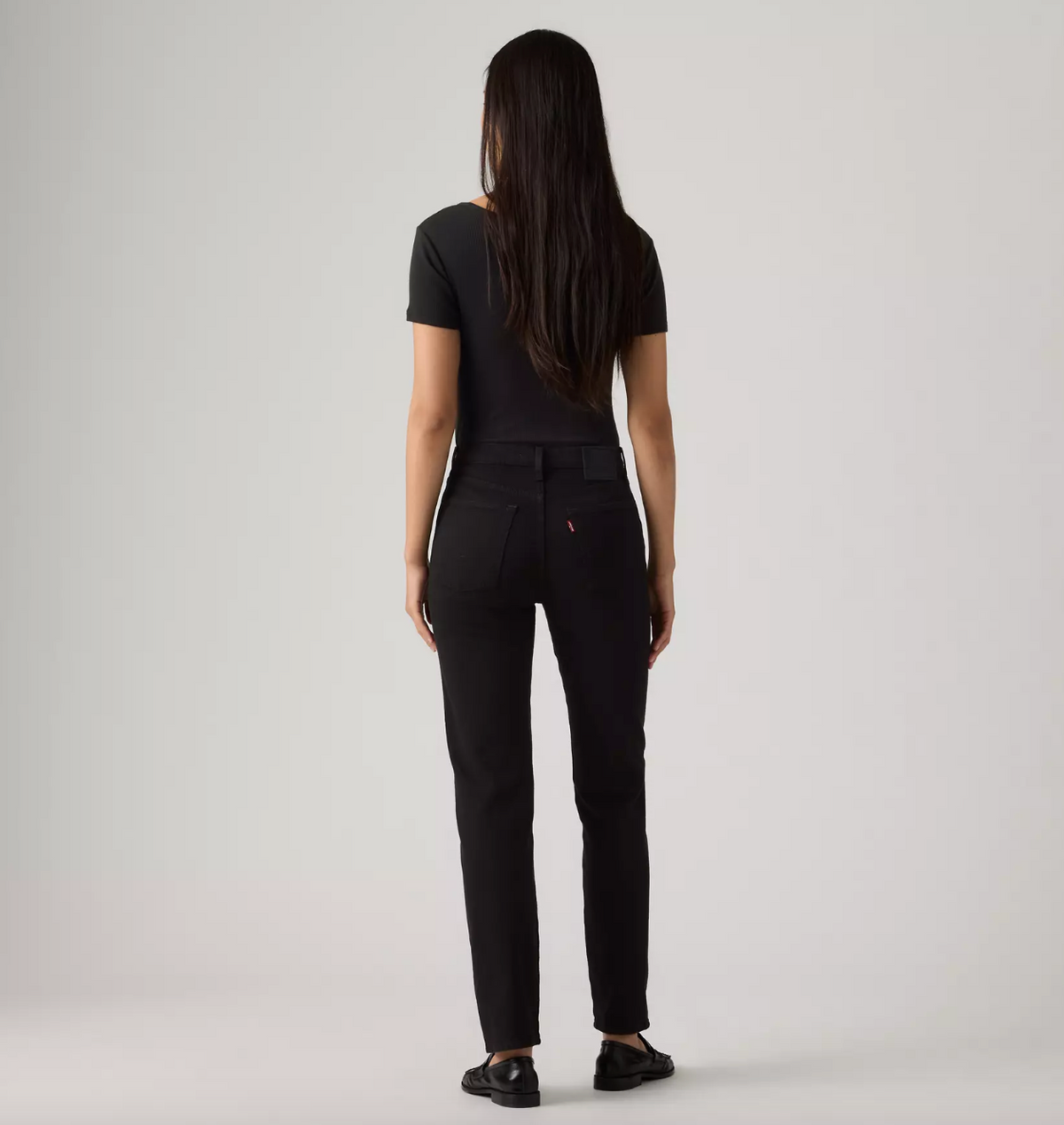 A woman with long dark hair is shown from behind wearing slim-fit black pants, a black short-sleeve top, and black shoes against a plain light gray studio background.