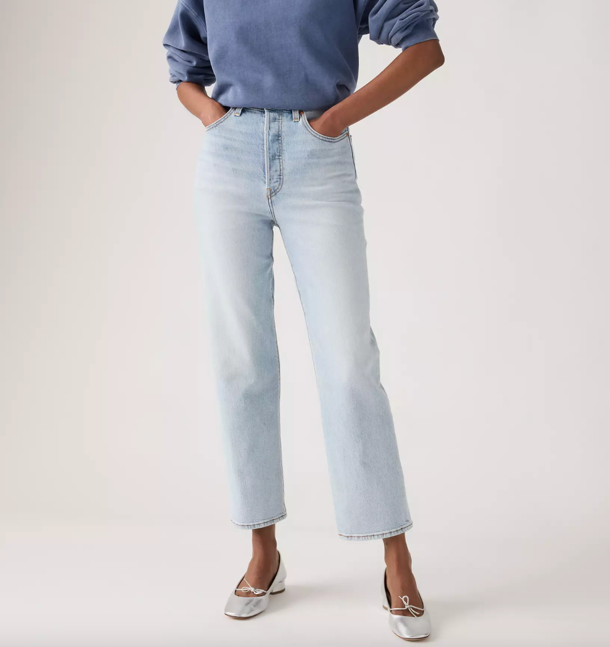 A model wearing light-wash light blue cropped jeans, a blue long-sleeve top, and silver flat shoes, standing against a plain off-white studio background.
