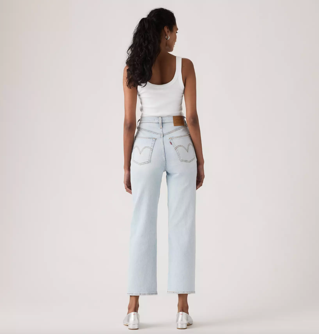 A woman is shown from behind wearing light-wash blue jeans with a brown leather waistband patch and silver pointed-toe heels, paired with a white tank top, against a plain off-white studio background.