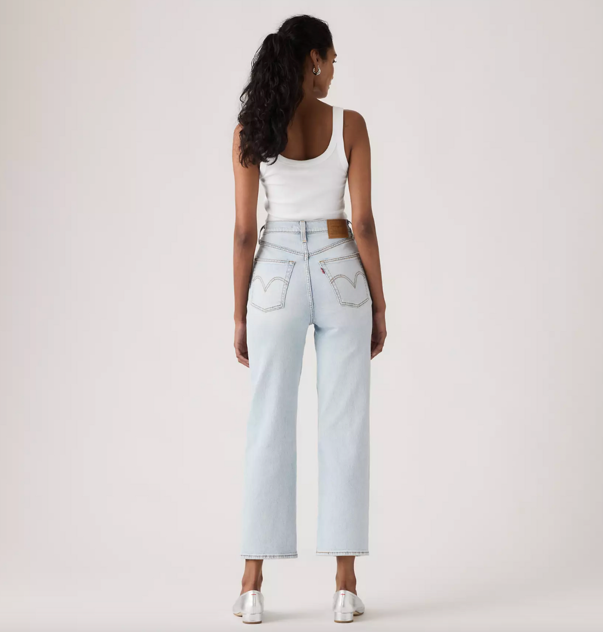 A woman is shown from behind wearing light-wash blue jeans with a brown leather waistband patch and silver pointed-toe heels, paired with a white tank top, against a plain off-white studio background.