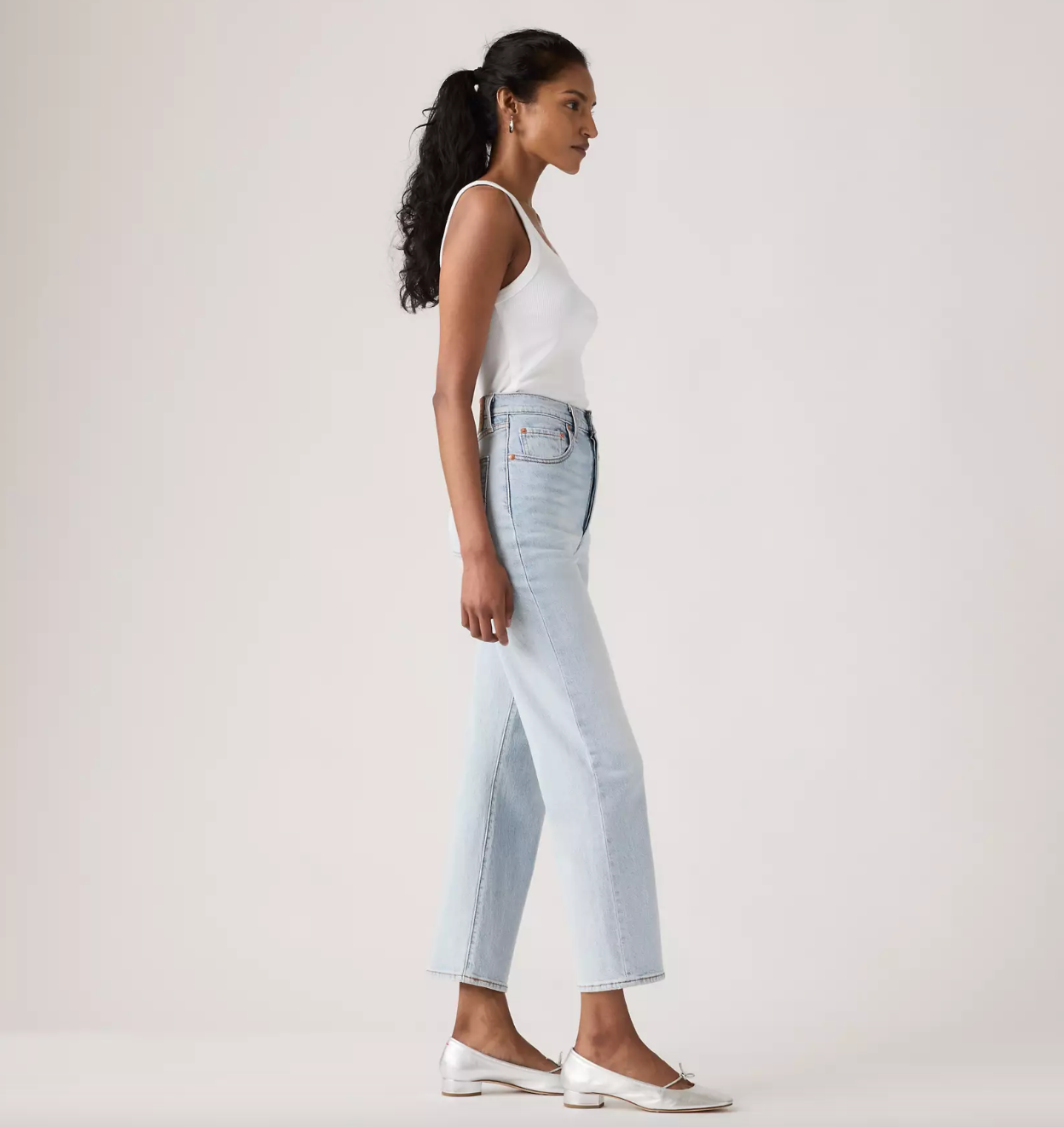 A woman in a white tank top and light-wash cropped jeans stands in profile against a plain light gray studio background, wearing white flats.