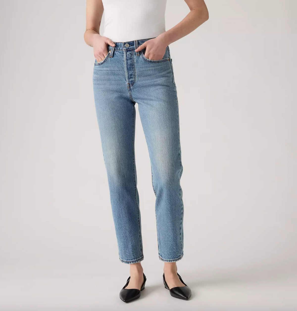 A model wearing light-wash blue straight-leg jeans, a white sleeveless top, and black pointed flats, standing against a light gray studio background.