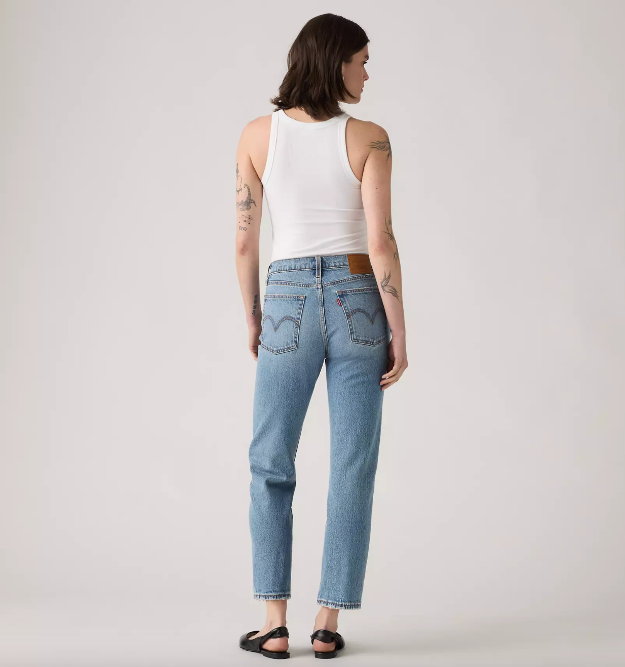 A model shown from the back wearing light-wash blue jeans, a white tank top, and black sandals against a plain light gray studio background.