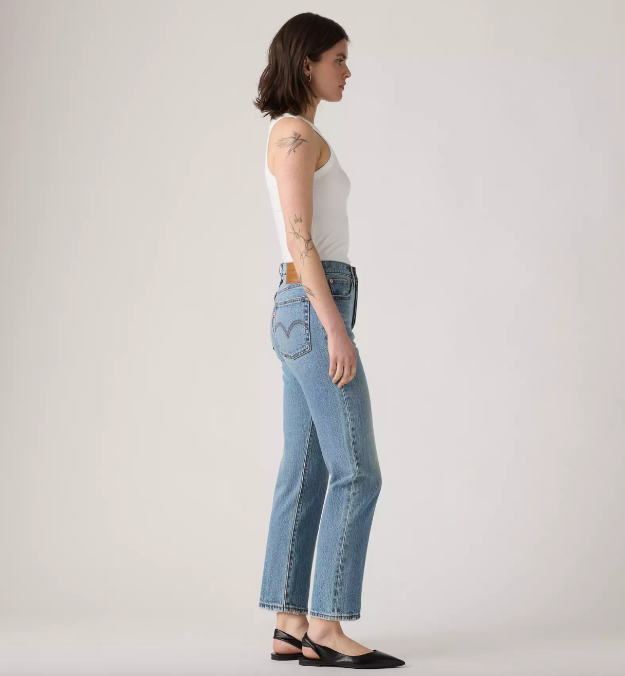A woman stands in profile wearing light-wash blue jeans, a white sleeveless top, and black flats against a plain light gray studio background.