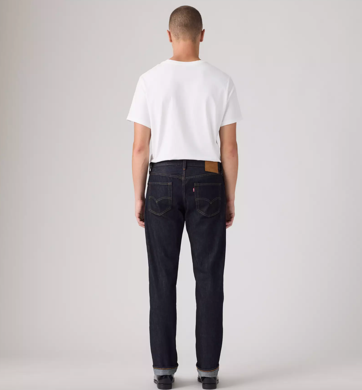A man viewed from behind wearing a white t-shirt and dark wash denim jeans with a brown leather patch on the back waistband, standing against a light gray studio background.