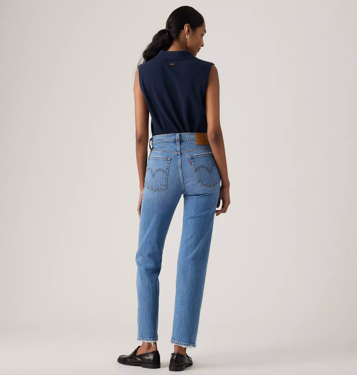 A model is shown from the back wearing a navy sleeveless collared top, light blue Levi's jeans with the brand's signature heart patch on the back pockets, and black leather loafers, against a plain light gray studio background.