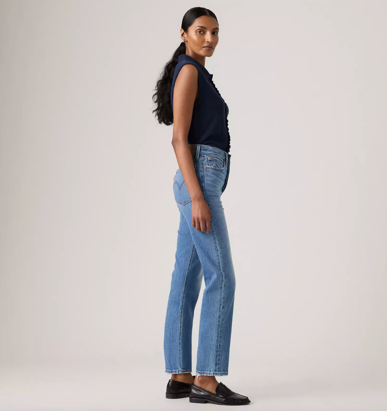 A woman modeling a pair of light-wash blue straight-leg jeans, paired with a navy sleeveless top and black loafers, standing in profile against a light gray studio background.