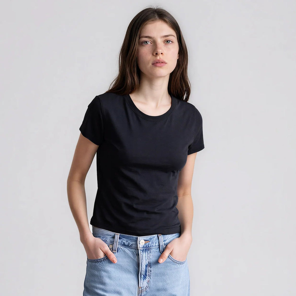 Model wearing It Is Well Cropped Tee in Black, front view against light grey background showing solid black lightweight slub jersey, clean crew neck, short fitted sleeves with slight cuff, and slightly cropped length in slim petite fit, styled with light wash high-rise jeans, hands in front pockets
