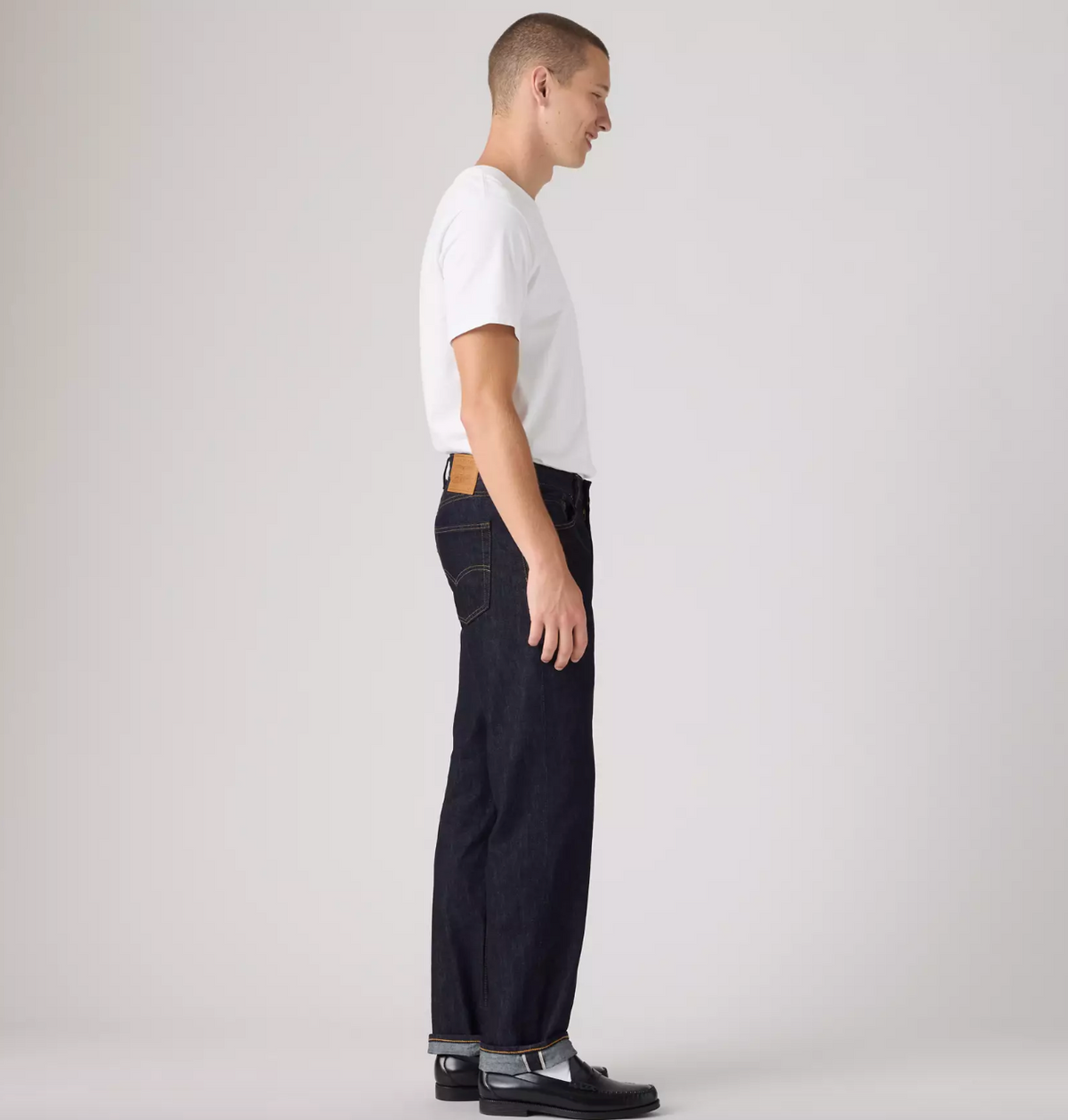 A man shown in profile wearing a white short-sleeve T-shirt, dark indigo jeans, and black leather shoes against a plain light gray studio background.