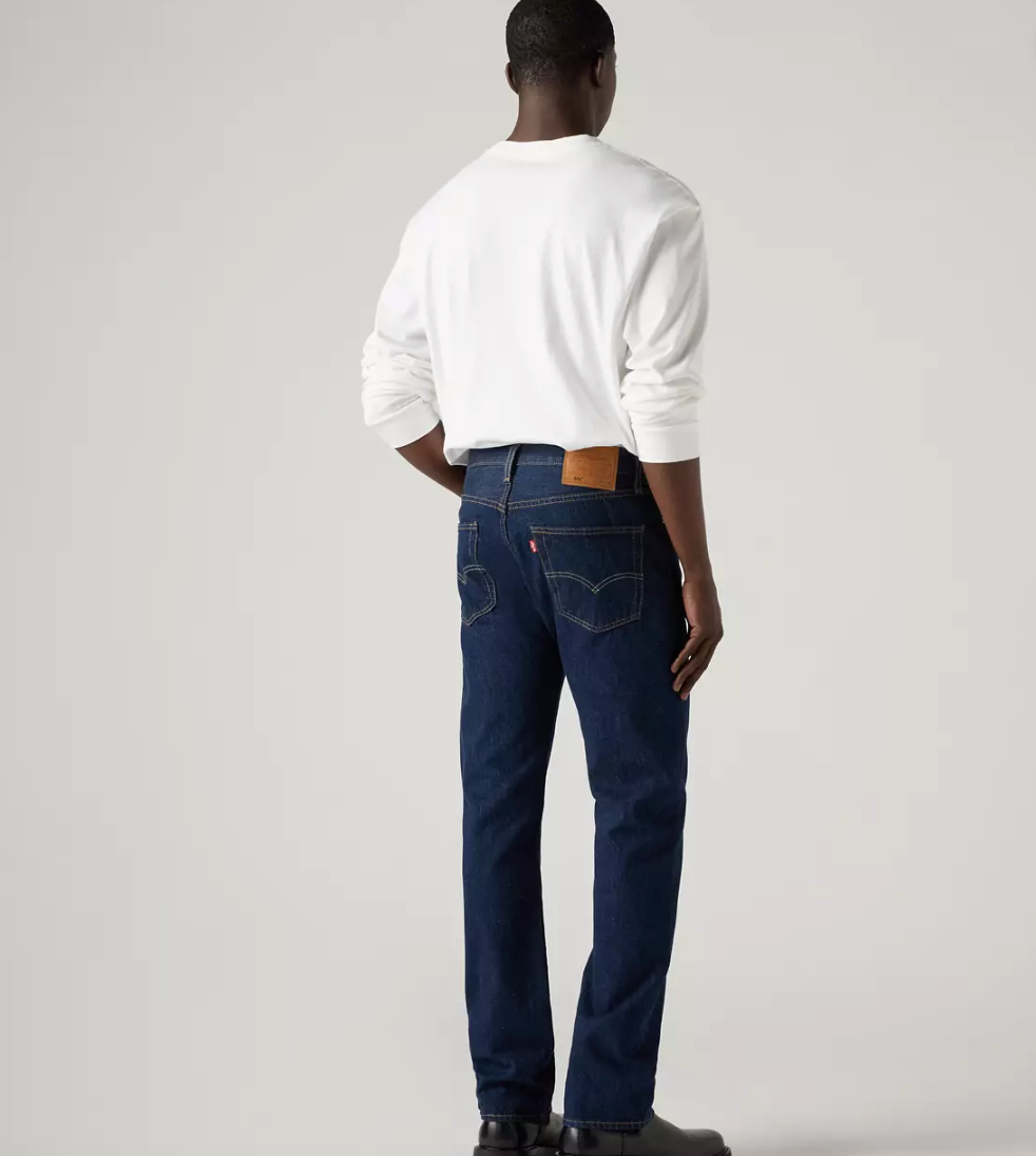 A man viewed from behind wearing a white long-sleeve crewneck shirt and dark blue Levi's jeans, standing against a light gray studio background.