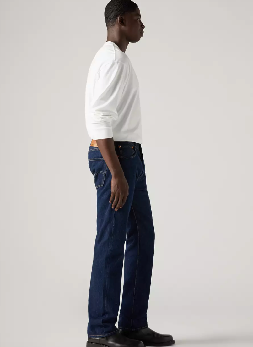 A man in profile wearing a white long-sleeve shirt and dark blue jeans, standing against a light gray studio background.