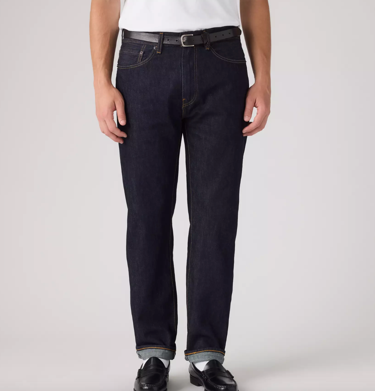 A person wearing dark indigo straight-leg jeans with a black belt, styled with a white shirt and black leather shoes, photographed against a light gray studio background.