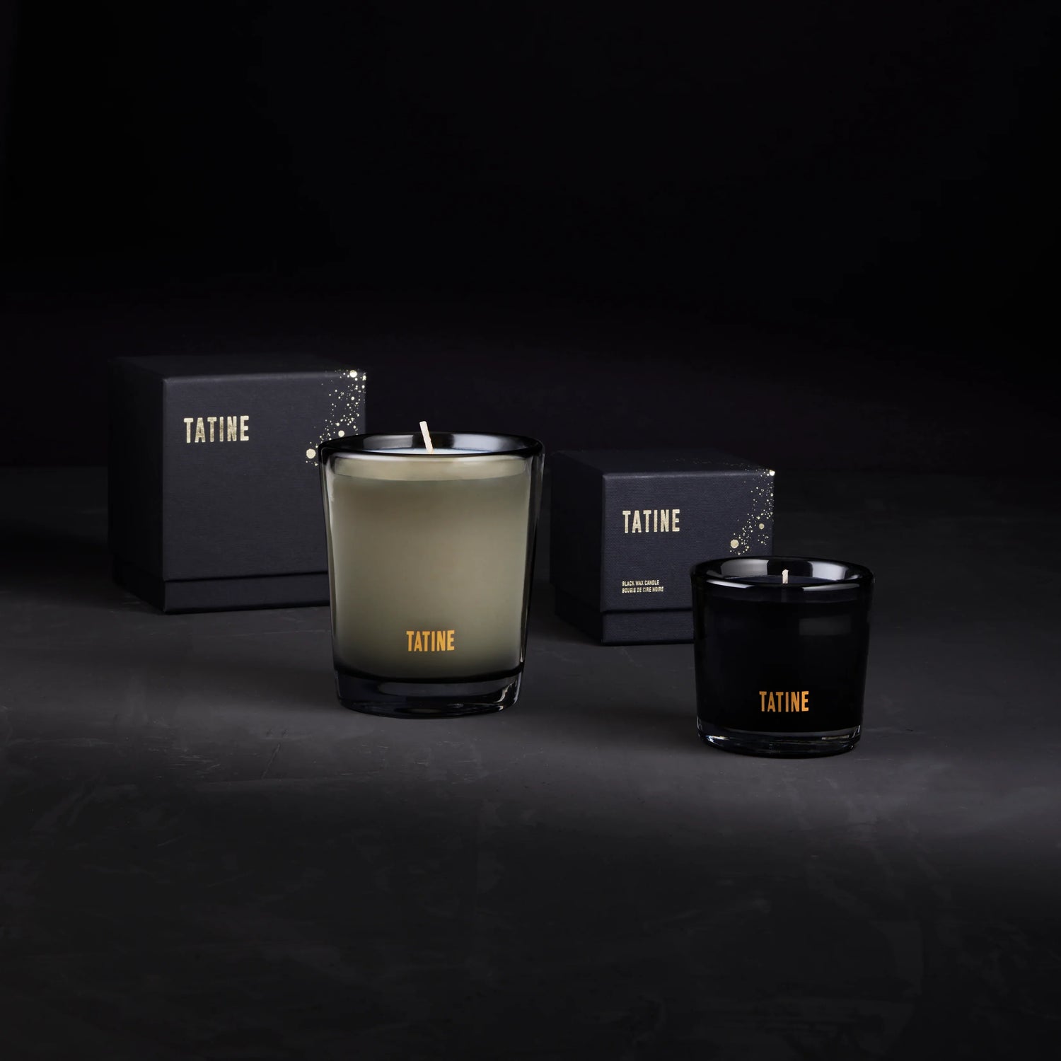 Two Tatine candles, one green and one black, with boxes in the background on a dark surface.