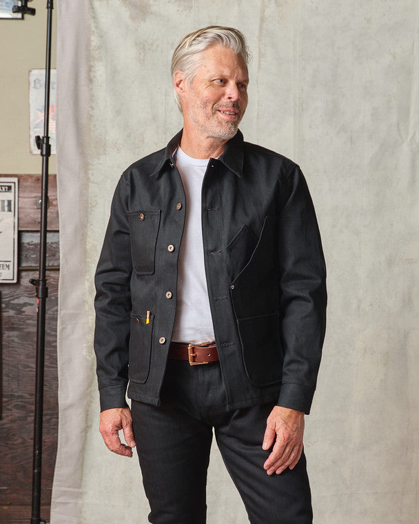 Model wearing the Tellason Coverall Jacket in Black Selvedge Denim open over a white t-shirt, styled with a brown leather belt and black pants, standing in a studio setting