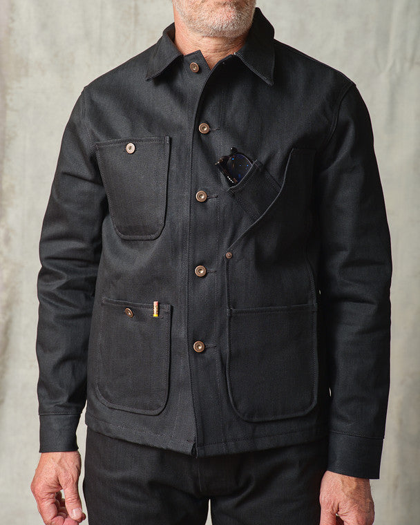 Close-up front view of the Tellason Coverall Jacket in Black Selvedge Denim, showing the point collar, copper button front, chest pocket with sunglasses, and lower utility pockets with Tellason label detail