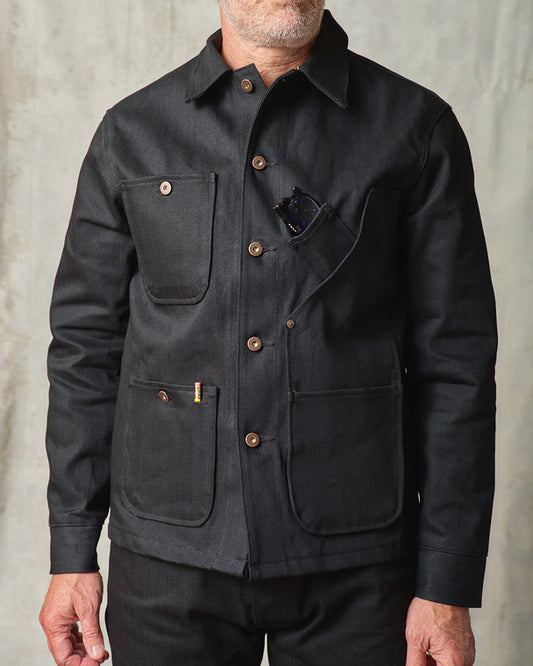 Close-up front view of the Tellason Coverall Jacket in Black Selvedge Denim, showing the point collar, copper button front, chest pocket with sunglasses, and lower utility pockets with Tellason label detail