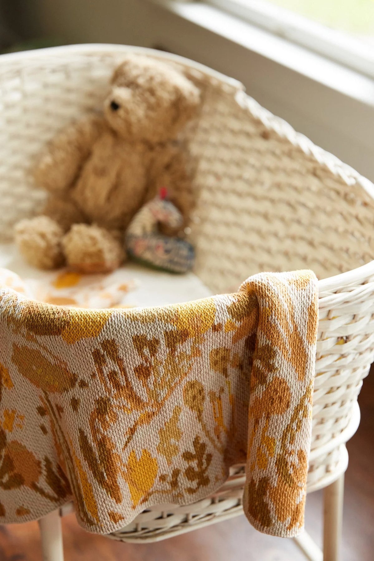 Poppies & Lotus Ocre blanket draped over a white woven bassinet, showing its golden floral knit pattern.
