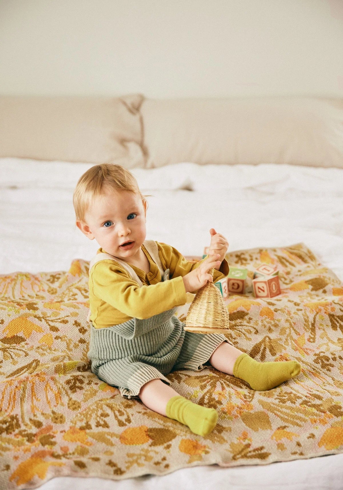 Child sitting on the Poppies & Lotus Ocre blanket spread across a bed, showing its golden and brown floral pattern.
