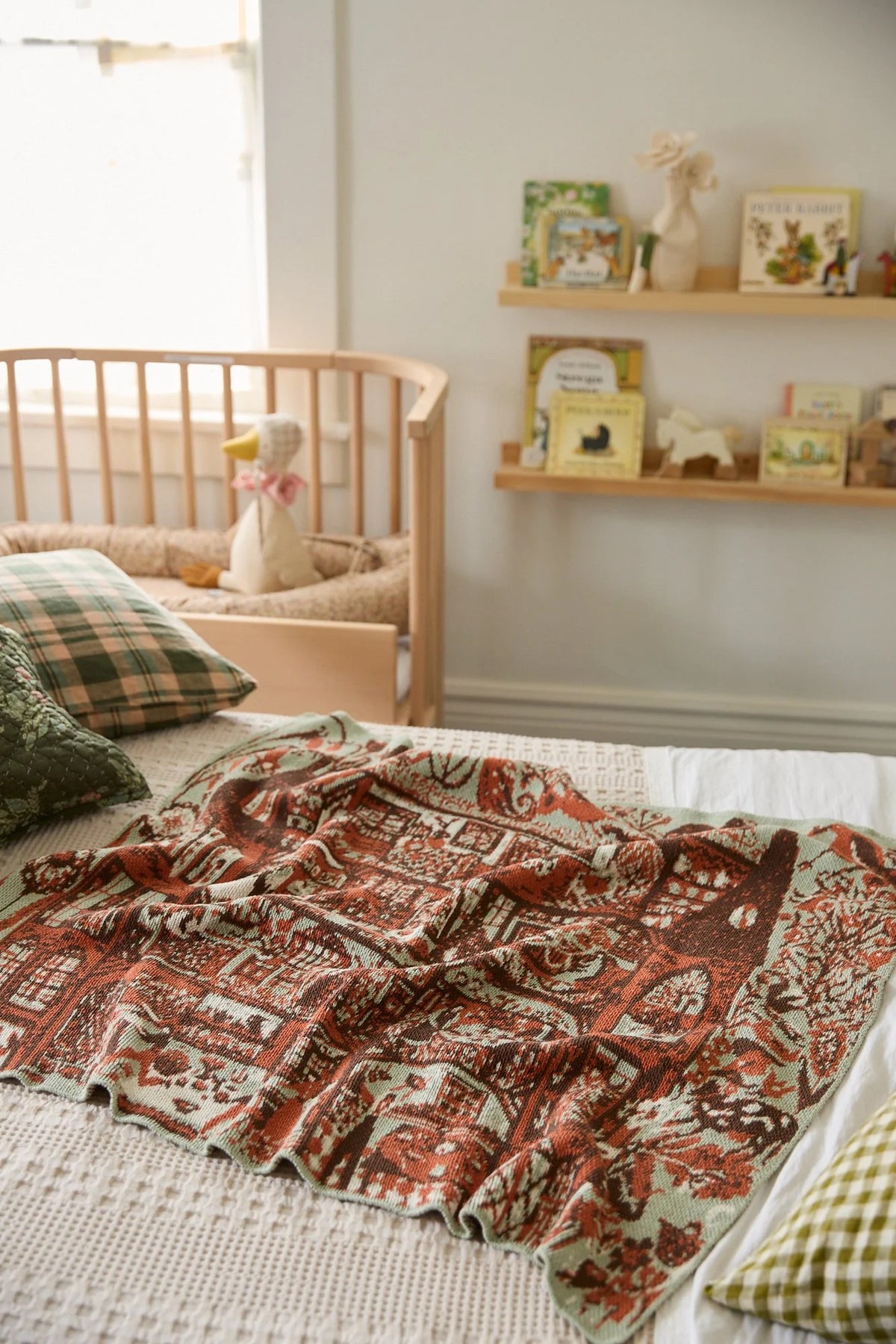 Swedish Farmhouse Sky Green blanket spread across a bed in a nursery, showing its red farmhouse pattern and pale green edge.
