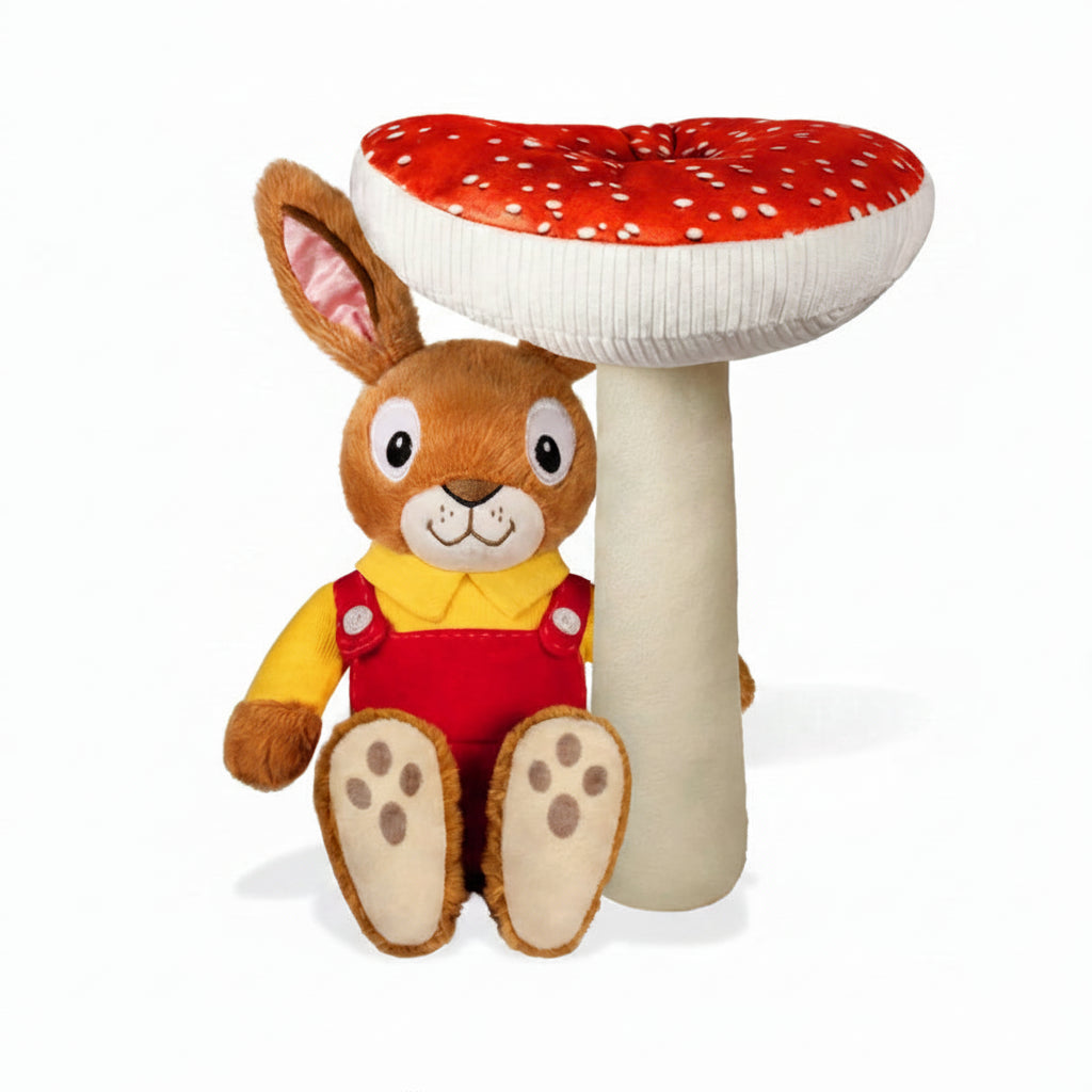 Nicholas bunny plush toy seated in front of a red-and-white spotted toadstool. The soft toy wears red overalls and a yellow shirt, with embroidered facial features, against a white background.
