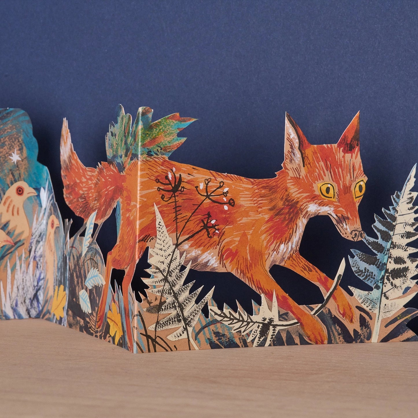 Close-up detail of the Three Fold Fox concertina card showing a vividly illustrated orange fox mid-stride through ferns and winter botanicals, with birds visible on the adjacent panel