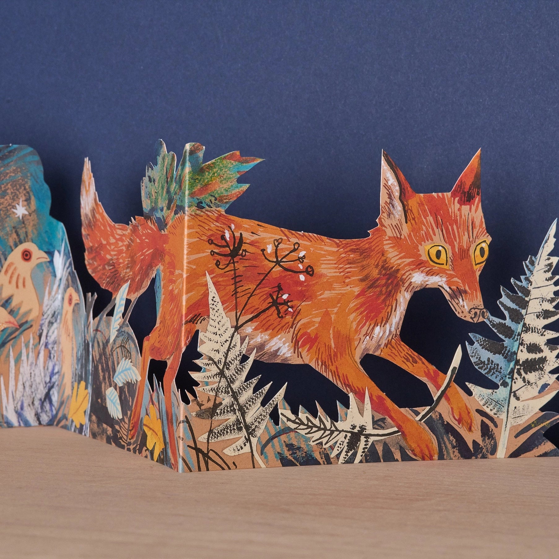 Close-up detail of the Three Fold Fox concertina card showing a vividly illustrated orange fox mid-stride through ferns and winter botanicals, with birds visible on the adjacent panel