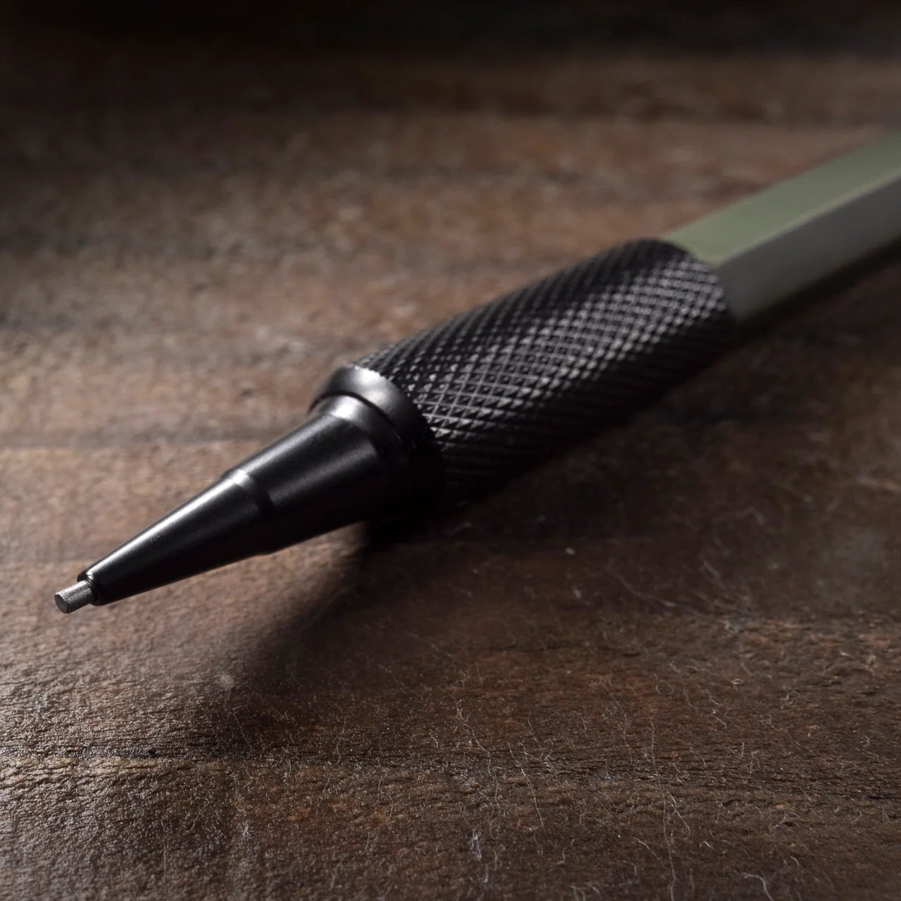 A close-up of a mechanical pencil with a textured black grip and a dark metallic tip, resting on a dark wooden surface.
