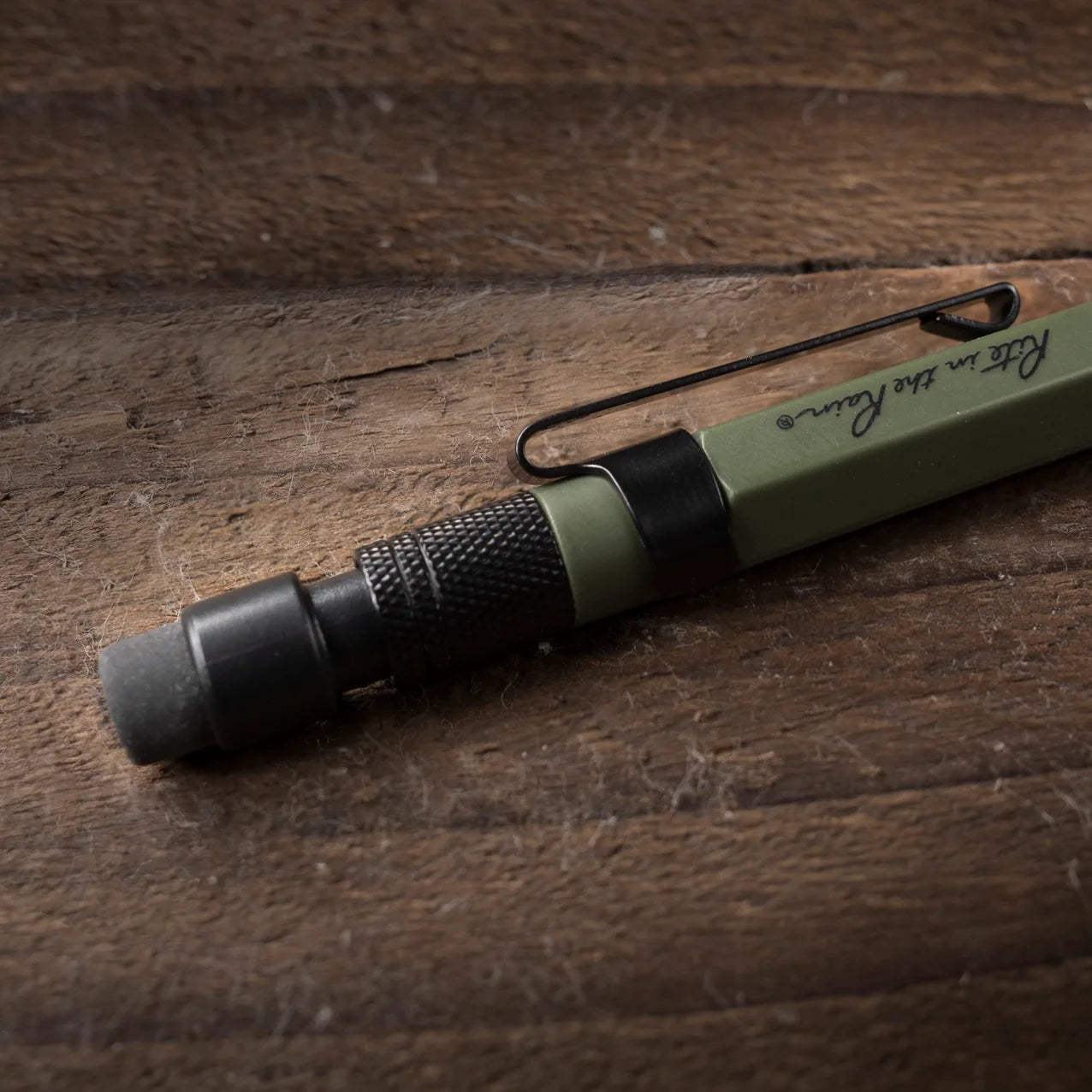 An olive green and black mechanical with a black pocket clip, resting diagonally on a rustic dark wood surface. The handle features a textured black grip and the words 'Rite in the Rain' printed in black script.