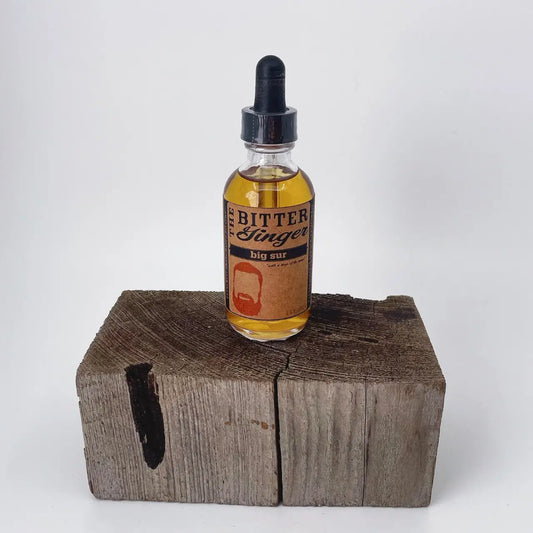 The Bitter Ginger Big Sur Bitters 2 oz glass dropper bottle with kraft paper label, displayed upright on a rustic weathered wood block against a white background