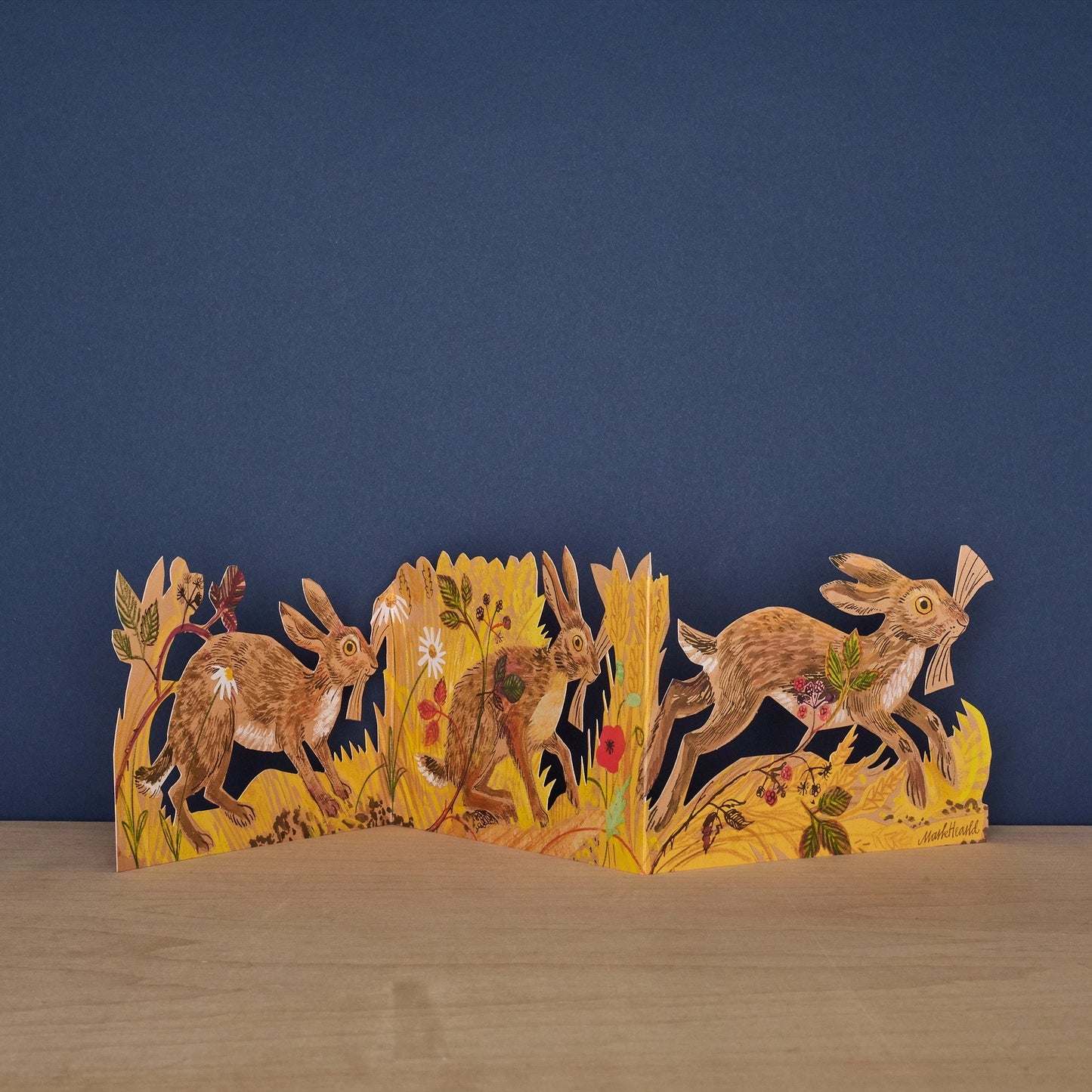 Hares Collage concertina card by Mark Hearld, fully extended showing three illustrated hares running through golden meadow grasses and wildflowers against a navy background