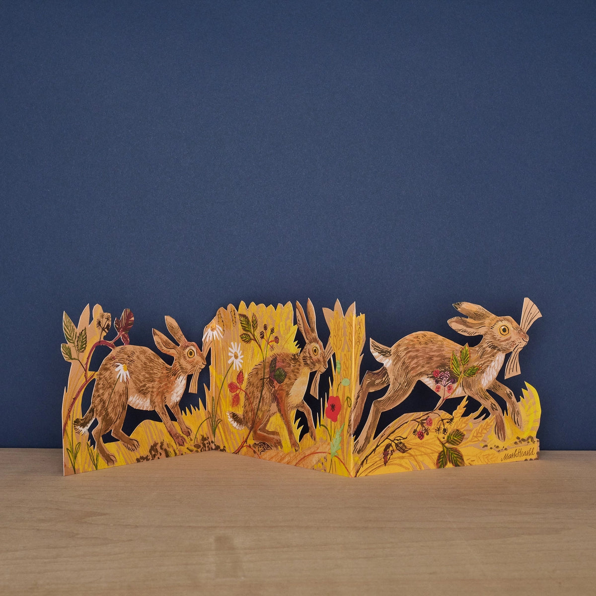 Hares Collage concertina card by Mark Hearld, fully extended showing three illustrated hares running through golden meadow grasses and wildflowers against a navy background
