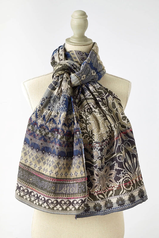 Létol Anoushka Scarf in Jean Trellis, draped and knotted on a linen dress form against a light grey background, showing a reversible Jacquard-woven organic cotton scarf — one side featuring a dense blue and oatmeal floral and trellis pattern with small repeated botanical motifs, the other showing large khaki and oatmeal Mughal medallion and palm tree scrollwork on a taupe ground — with the woven Létol label at the lower center and a pink accent selvedge visible at the hem
